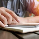 A close-up view of a hand interacting with a smartphone and notebook on a desk.