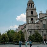 People walking near a historic architectural landmark with a prominent bell tower under a clear blue sky.