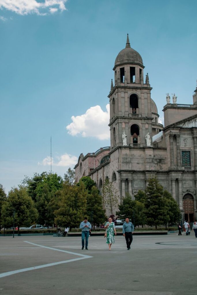 People walking near a historic architectural landmark with a prominent bell tower under a clear blue sky.